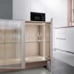 Modern kitchen cabinet with ribbed glass door open, revealing lit interior with three wine glasses on a shelf. Sleek white cabinetry and microwave in background.