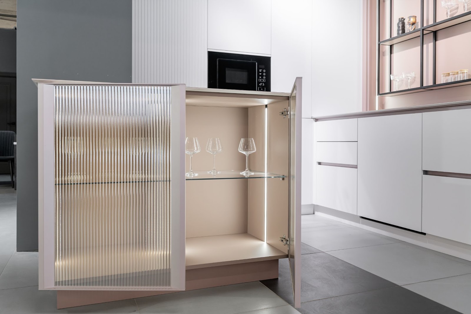 Modern kitchen cabinet with ribbed glass door open, revealing lit interior with three wine glasses on a shelf. Sleek white cabinetry and microwave in background.