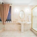Luxurious bathroom with marble walls, gold fixtures, pedestal sink, oval mirror, glass shower, pink curtains, and potted plants on window sill.