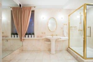 Luxurious bathroom with marble walls, gold fixtures, pedestal sink, oval mirror, glass shower, pink curtains, and potted plants on window sill.