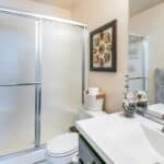 Modern bathroom with frosted glass shower doors,white toilet,single sink vanity with silver faucet,gray mosaic tile backsplash,and blue-gray towels.Decor includes seashell bowl and floral wall art.
