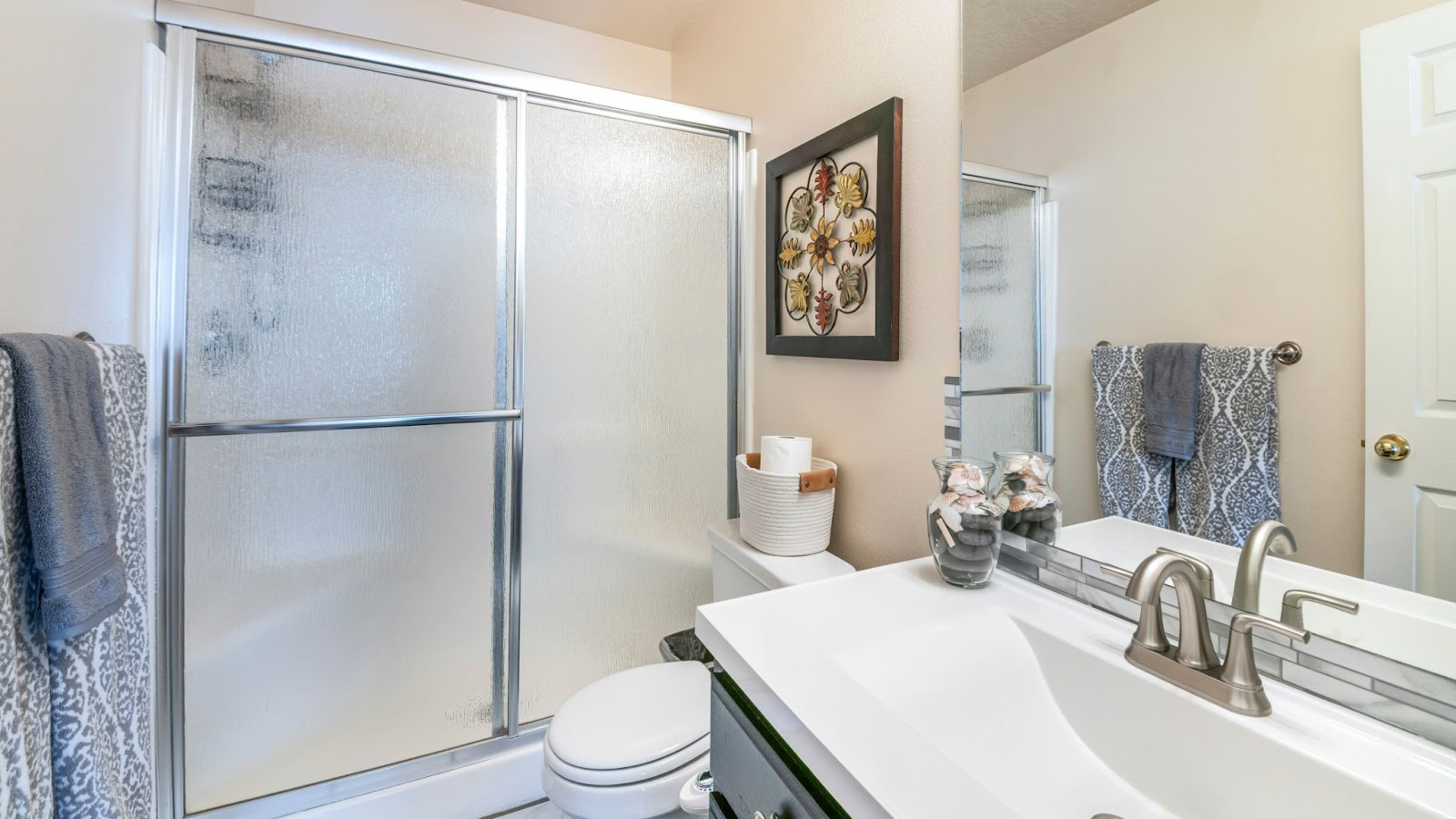 Modern bathroom with frosted glass shower doors,white toilet,single sink vanity with silver faucet,gray mosaic tile backsplash,and blue-gray towels.Decor includes seashell bowl and floral wall art.